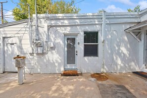 A white single-story building with a front door, a window with a shutter, and a small porch area. - Delightful 1BR 1BA Cozy Flat (Fort Worth)