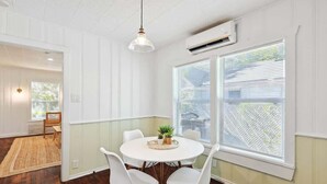 Een eetruimte met een ronde tafel, witte stoelen, een hanglamp en grote ramen met jaloezieën.