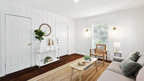 Een kamer met witte lambrisering, een houten vloer, een bank, een stoel, een bijzettafeltje en een spiegel.