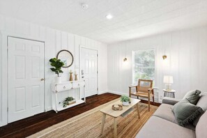 Une pièce aux murs recouverts de panneaux blancs, avec un plancher en bois, un canapé, une chaise, une petite table et un miroir.