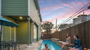 Un jardin à l’arrière d’une maison, avec une piscine, une clôture en bois et un espace patio aménagé avec des chaises et un parasol.