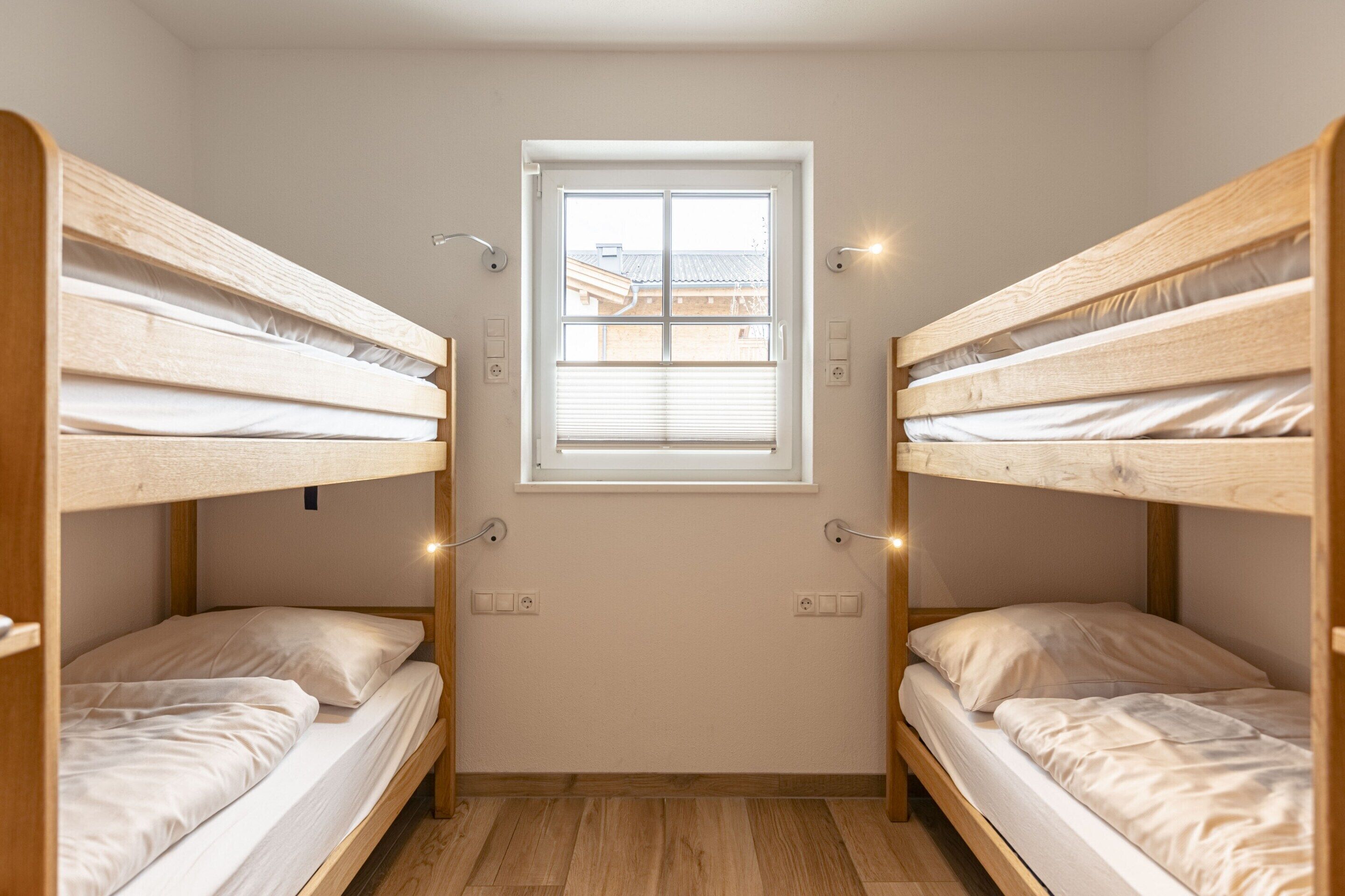 Quarto pequeno e bem iluminado com duas camas beliche, janela com persianas e vista para prédios.
