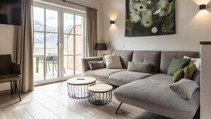 A modern living room with a large sofa, two round side tables, a flat-screen TV, and a large window with a view of mountains. - Superior Ferienhaus # 2B mit Sauna (Uttendorf)
