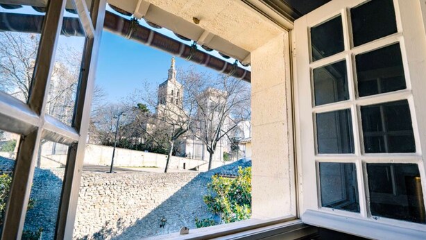 Una ventana con vista a un muro de piedra, un edificio con torre y cielo despejado.