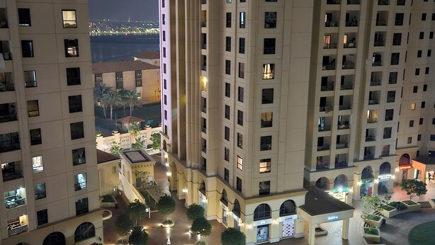 A high-rise residential building with multiple balconies, a central courtyard with trees and seating areas, and a view of the sea in the background. - SEASIDE HOSTELS (Dubai)