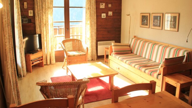 A cozy living room with a wooden floor, a striped sofa, a wicker chair, a wooden table, and a television on a stand.