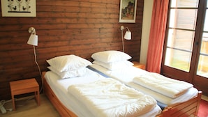 A double bed with white linens in a room with wooden panel walls, two wall-mounted lamps, and a window with curtains.