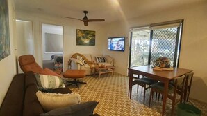 Un soggiorno con divano, poltrona e tavolo da pranzo con sedie. Si nota una ventola a soffitto, un televisore fissato alla parete e una finestra con vista.