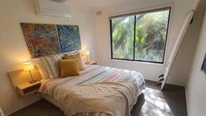 Una camera da letto con un letto, due lampade sui comodini, una finestra vista alberi e due quadri astratti alle pareti.