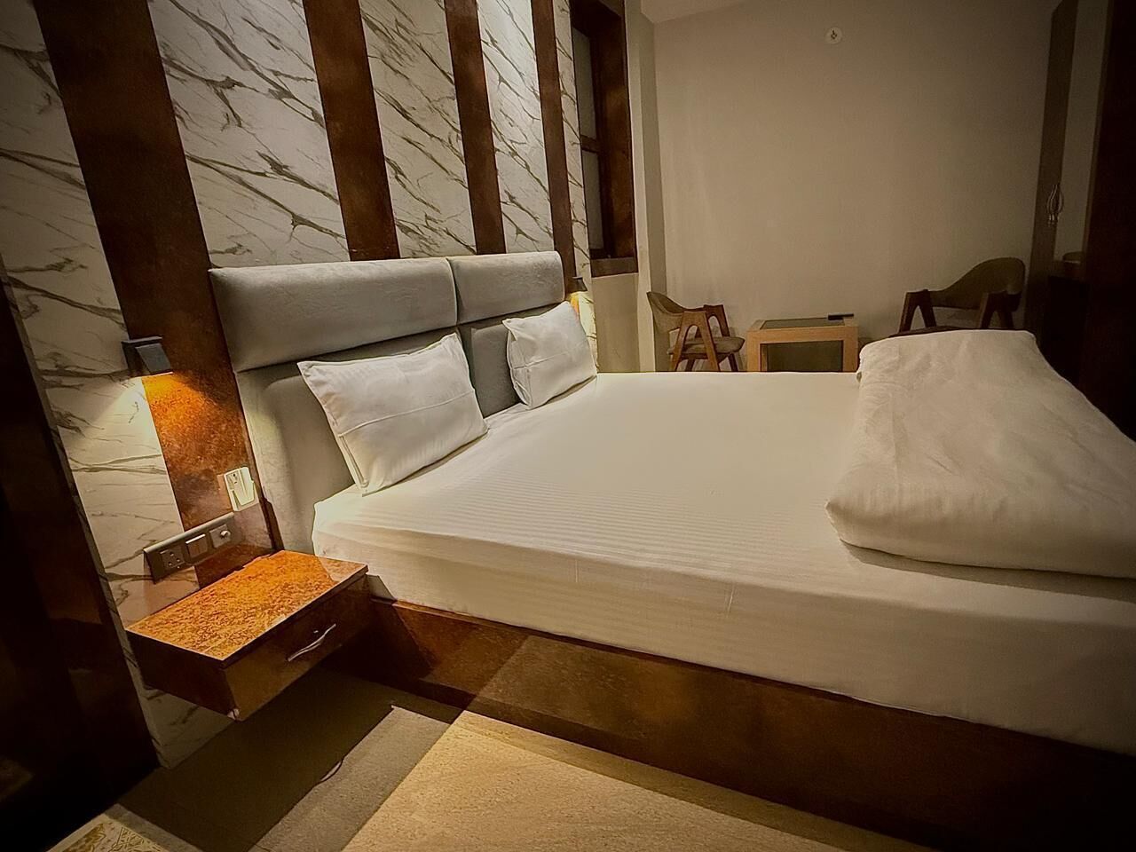 Una cama bien hecha con cabecera, una mesita de noche con lámpara y una silla en una habitación con pared de diseño marmolado.