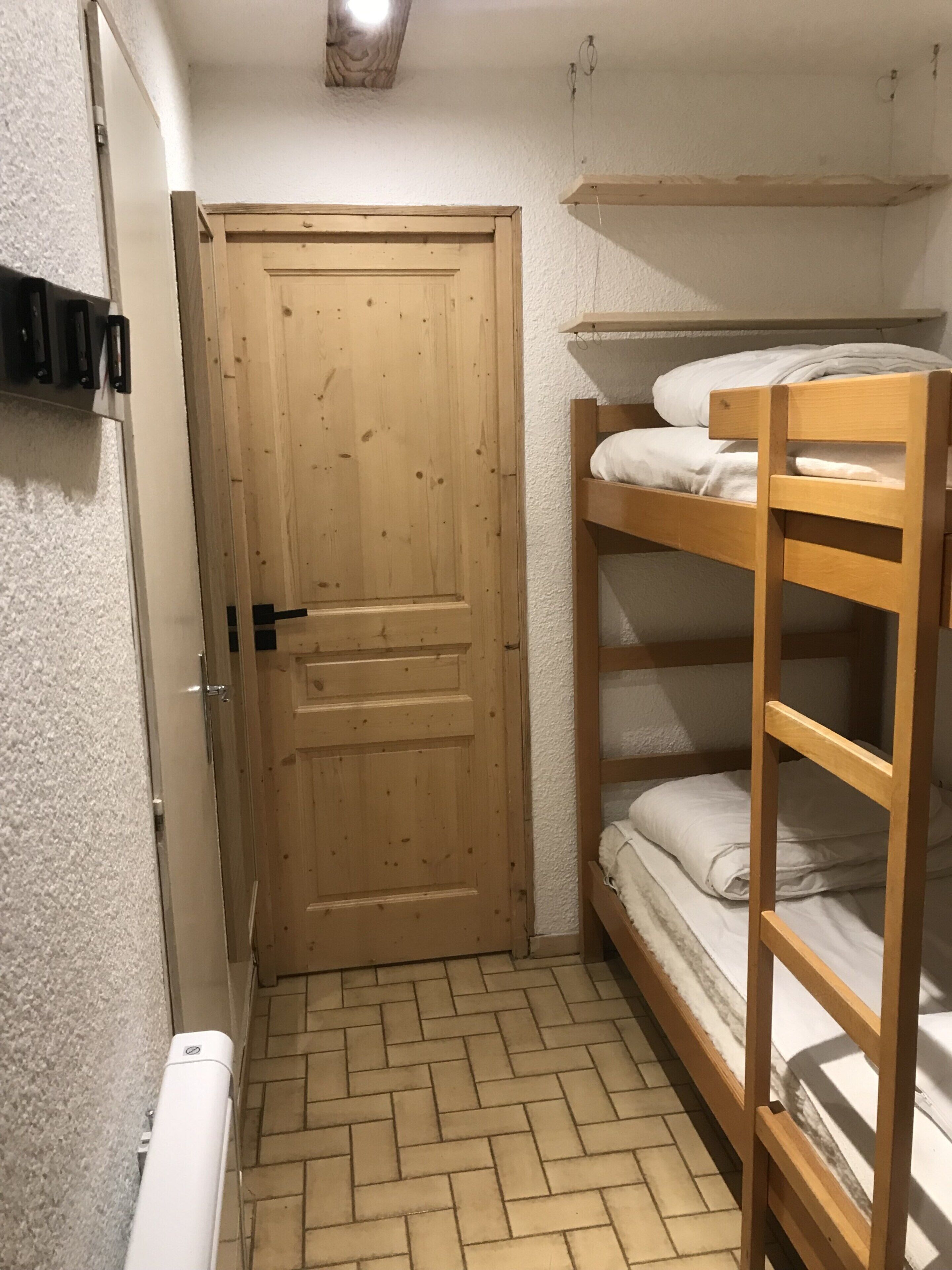 A small, narrow room with a wooden door, a bunk bed, and a radiator on the floor.