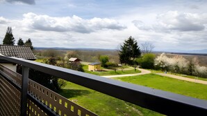 Un balcón con vista a un paisaje verde, una pequeña estructura de madera y montañas al fondo.