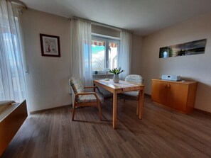 Een kamer met een houten vloer, een eettafel met stoelen, een radiator, een raam met gordijnen, een kast en een schilderij aan de muur.