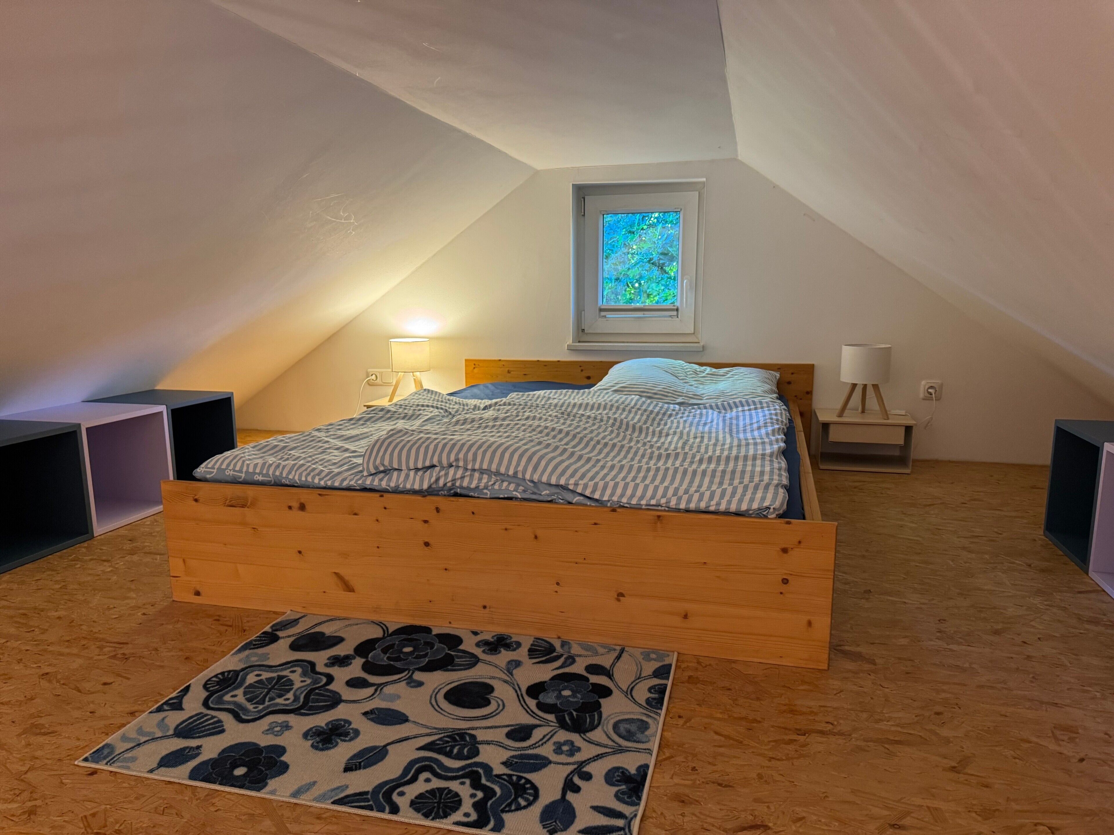 Una camera da letto con soffitto spiovente, un letto con struttura in legno, una finestra con vista e un comodino con lampada.
