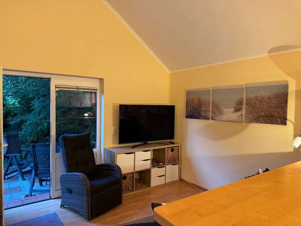A living room with a TV, a chair, a table, and a sliding glass door leading to an outdoor area with chairs.