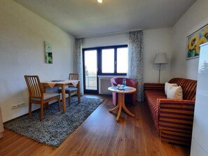 Una habitación con una mesa de comedor, sillas, un sofá, una mesita redonda con una botella, un radiador, una ventana con cortinas y una lámpara.