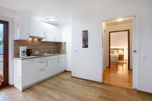 Una cocina moderna con armarios blancos, una encimera de madera y una cafetera. Se ve un pasillo que conduce a un dormitorio con cama y mesita de noche.