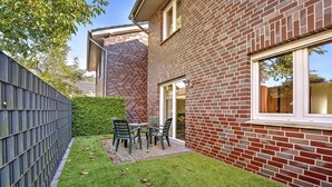Een bakstenen huis met een klein gazon, een patio tafel met stoelen en een metalen hek.