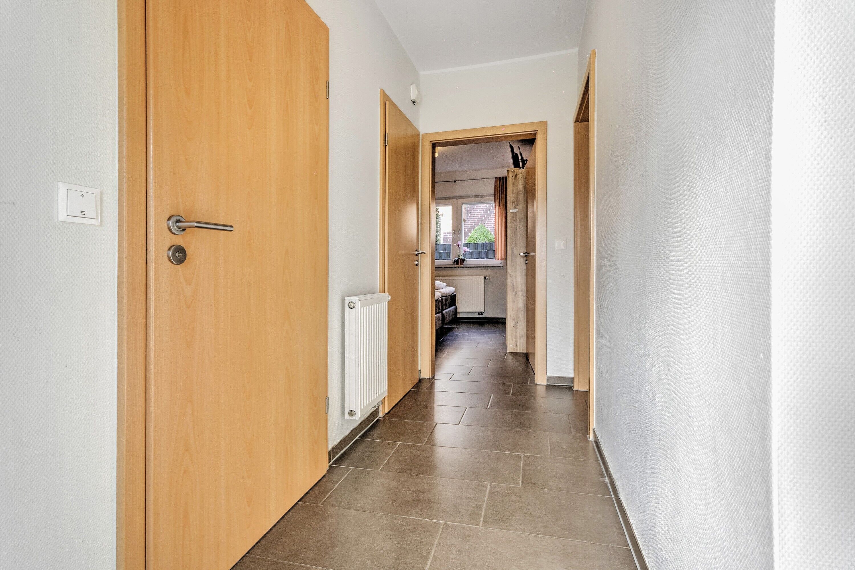 A hallway with a wooden door, a radiator, and a view into a kitchen area.