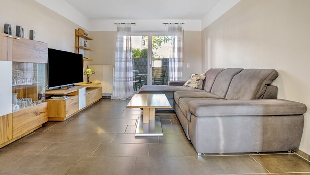 Een moderne woonkamer met een flatscreen televisie, houten meubels, een grijze bank en een schuifpui naar buiten.