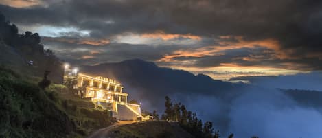 A modern building with a staircase leading to the entrance, surrounded by lush greenery and a mountainous landscape.