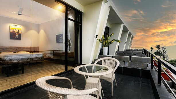 Quarto de hotel moderno com parede de vidro, cama, área de estar com cadeiras de vime e varanda com vista para o pôr do sol.