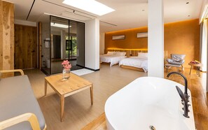 A modern hotel room with a large bathtub, two beds, a wooden table, and a glass door leading to a bathroom.