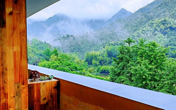 A view from a balcony with a wooden railing, overlooking a lush green landscape with dense foliage and mist-covered mountains in the background.