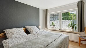 A bedroom with a bed, two pillows, a nightstand, a lamp, a window with curtains, and a potted plant.