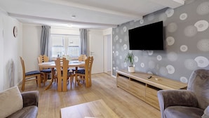A living room with a dining area, a flat-screen TV mounted on the wall, and a potted plant on the entertainment unit.