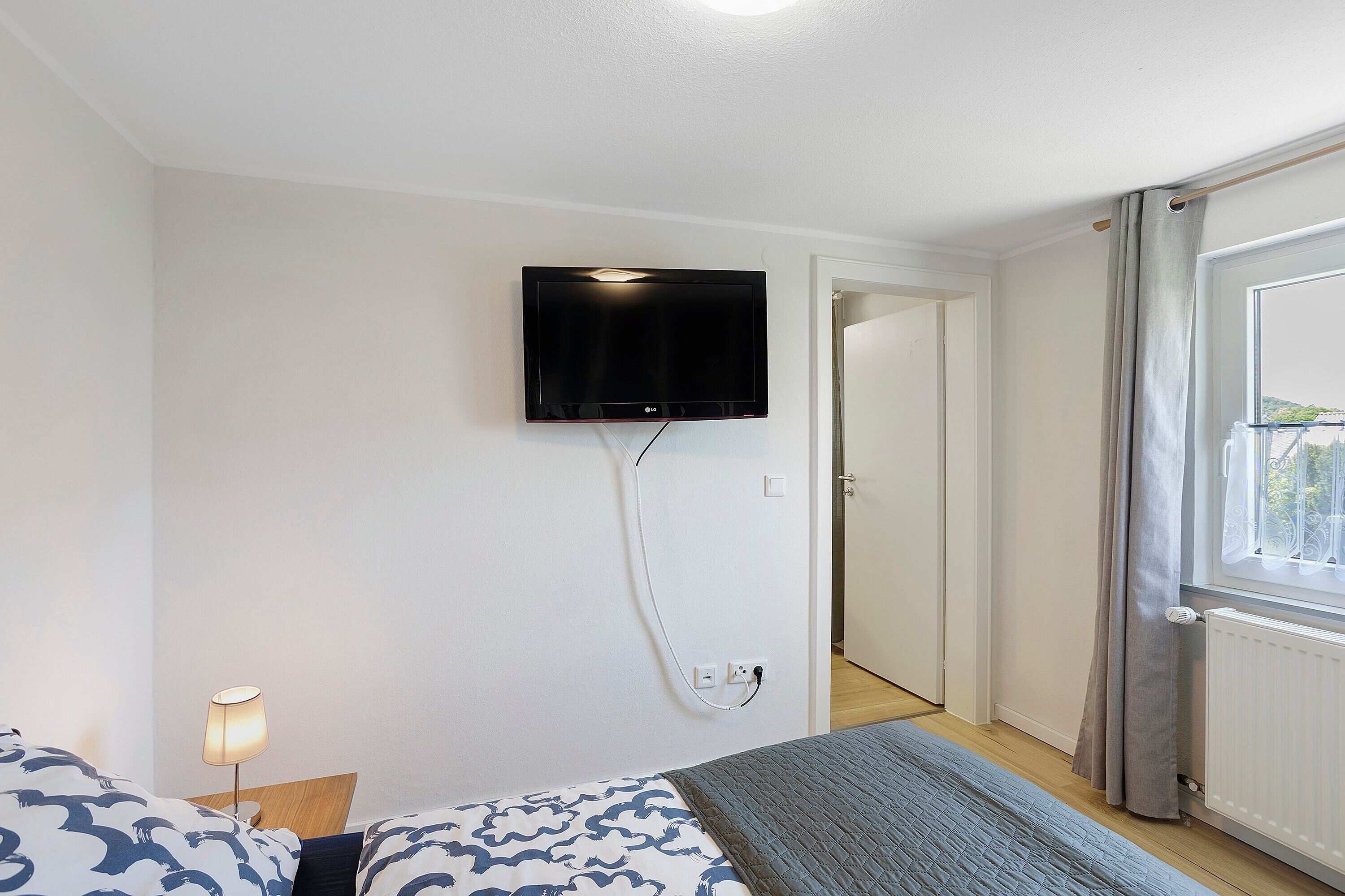 Un dormitorio con cama, televisión instalada en la pared, lámpara de noche y ventana con cortinas.