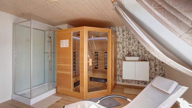 A room with a glass shower enclosure, a wooden sauna, a white sink, and a reclining chair.