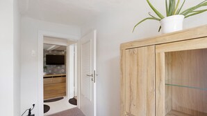 A hallway with a wooden cabinet, a potted plant, and a door leading to a room with a TV and a patterned rug.