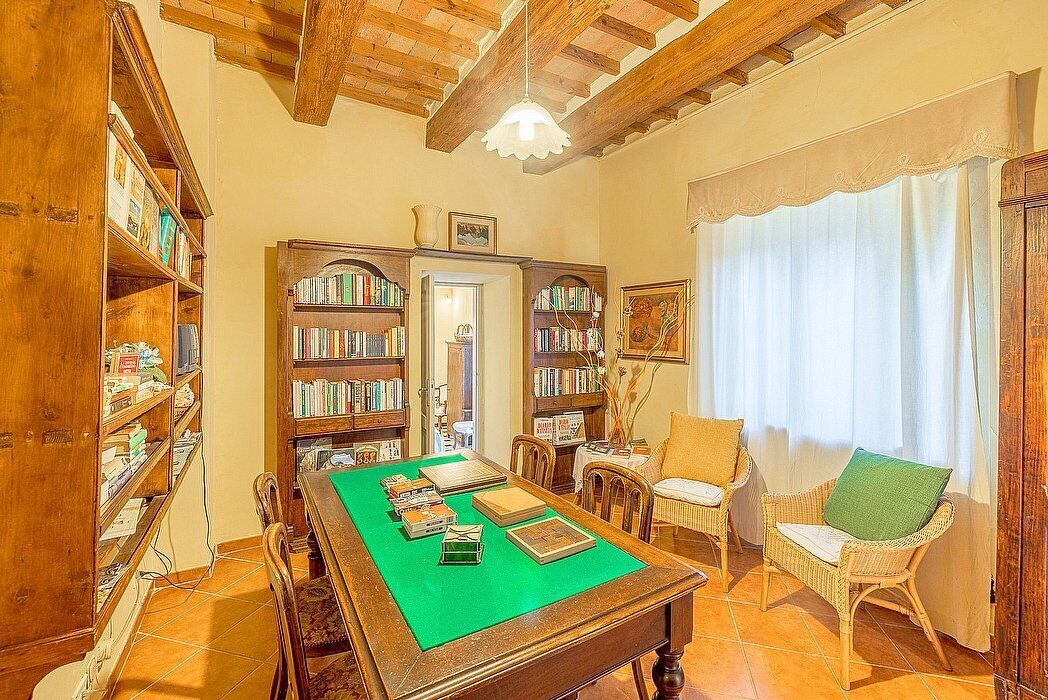 A room with a green table, wooden bookshelves, wicker chairs, and a ceiling with exposed beams.