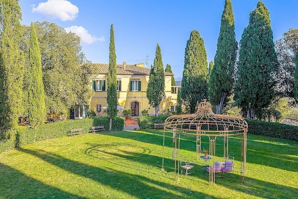 Une grande maison avec un jardin, un belvédère et de grands arbres.