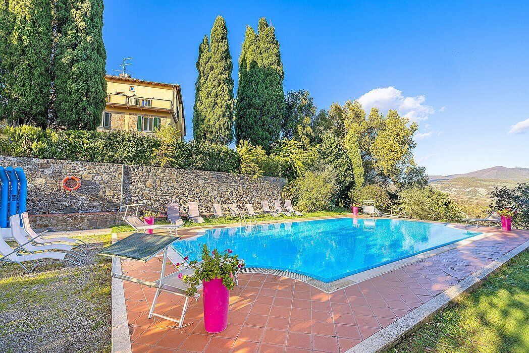 A swimming pool with lounge chairs, a stone wall, and tall trees.