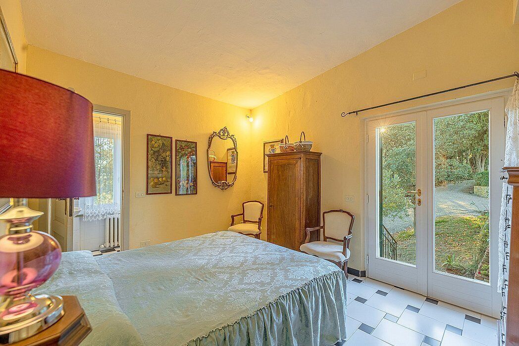 Un dormitorio con una cama, una lámpara, un armario, dos sillas, un espejo y un ventanal corredero que da a un jardín.