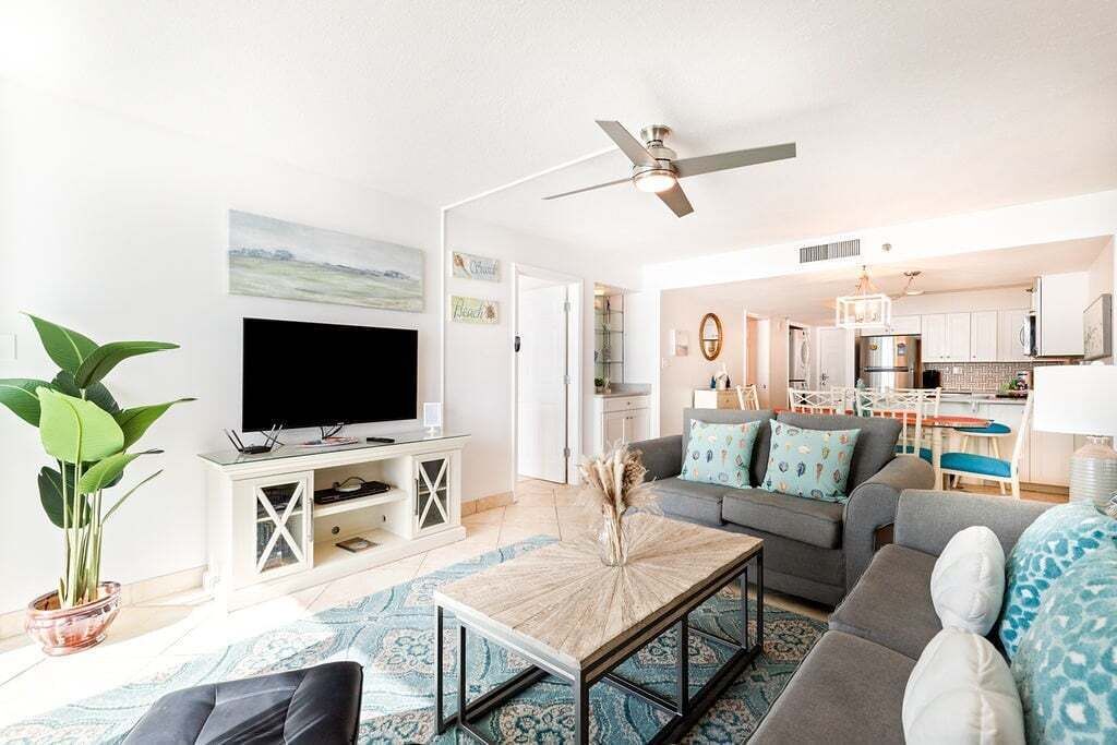 A living room with a flat-screen TV, a ceiling fan, a coffee table, and a sofa. There is a kitchen in the background.
