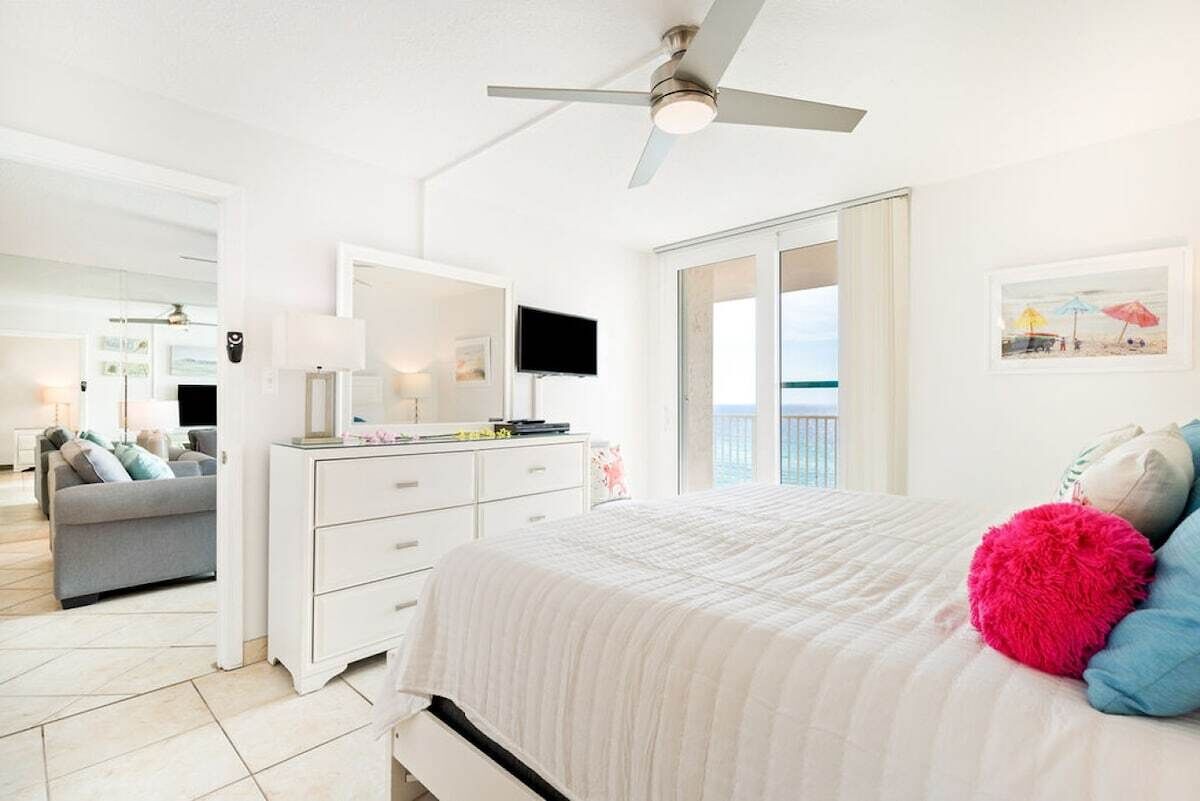 A bright bedroom with a bed, a ceiling fan, a TV, and a balcony with a view.