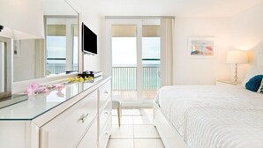 Habitación de hotel con cama, cómoda, espejo, televisor y balcón con vista al océano.