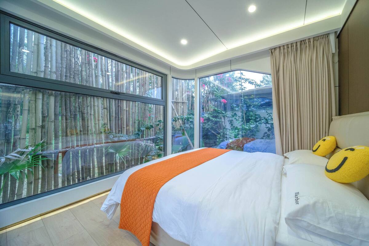 A modern hotel room with a large bed, a view of bamboo outside, and a ceiling with recessed lighting.