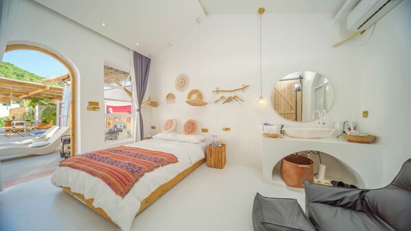 Un dormitorio con cama, tocador con espejo, un taburete de madera y vista al exterior.