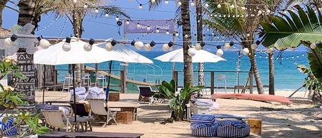 Un café frente a la playa con mesas al aire libre, palmeras y luces colgantes.