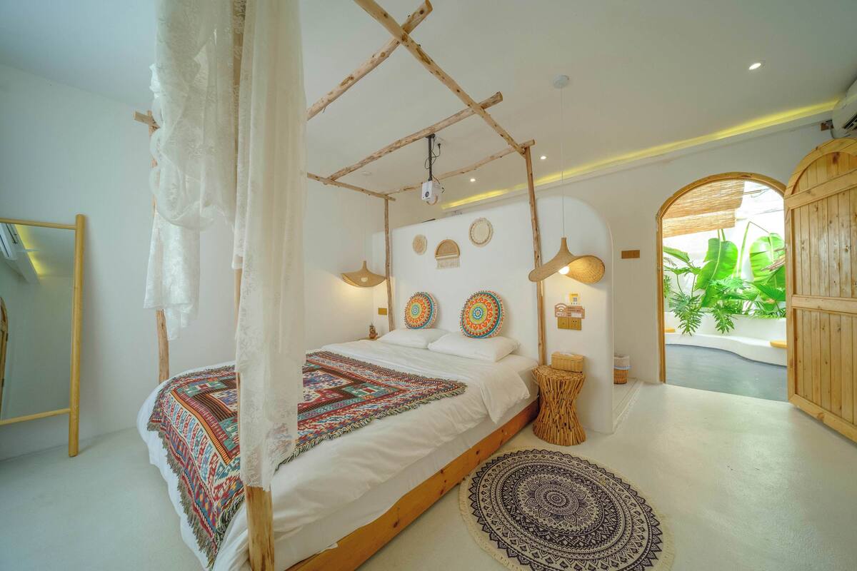 A bedroom with a canopy bed, a large mirror, a wooden door, and a view of greenery outside.