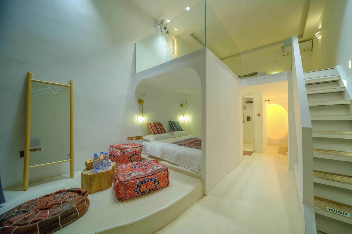 A hotel room with a bed, a small table with water bottles, a mirror, and a staircase.