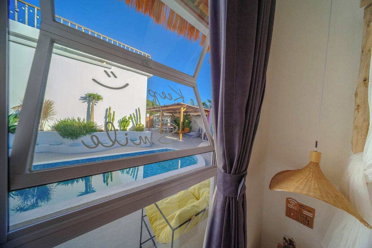 A room with a large window overlooking a pool area, a bed with a white comforter, and a wooden ceiling fan.