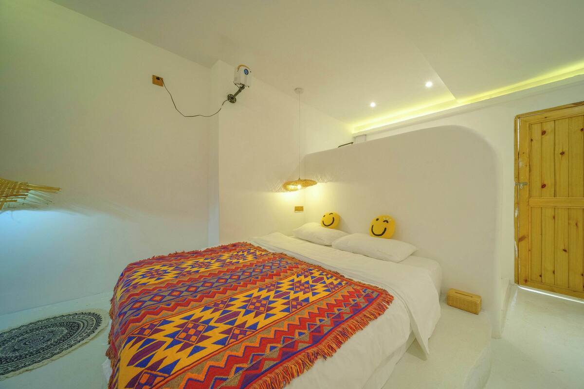A bedroom with a bed, a wall-mounted clock, a decorative wall hanging, and a wooden door.