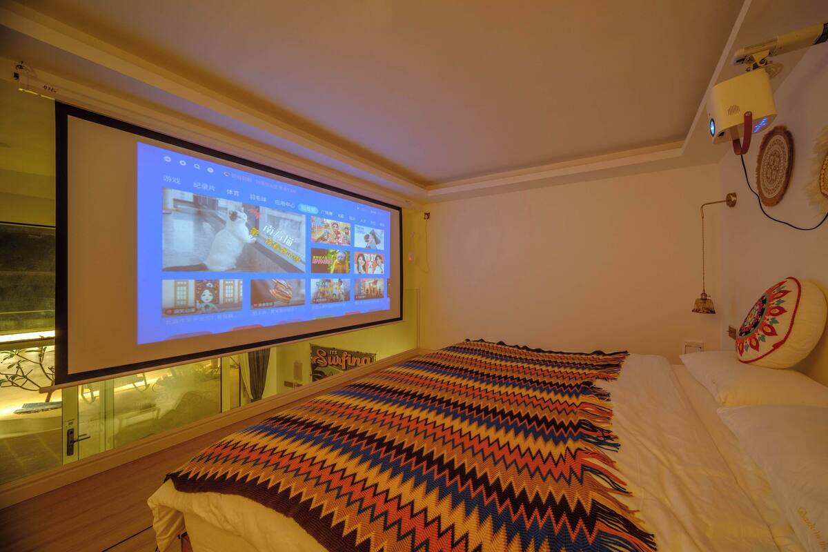 A hotel room with a bed, a projector screen displaying a webpage, a window with a view of the outside, and a decorative wall hanging.