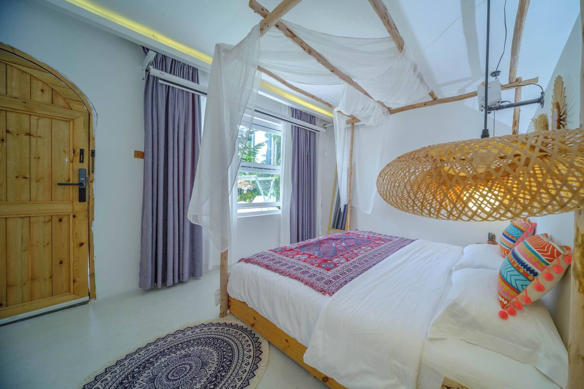 A bedroom with a bed, a wooden door, a window with curtains, a woven lamp, and a patterned rug.