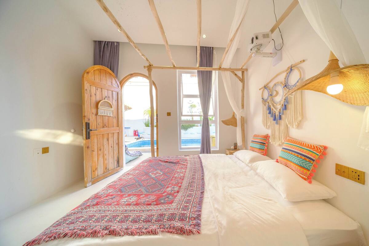 A bedroom with a large bed, a wooden door, a view of a swimming pool, and a macrame wall hanging.
